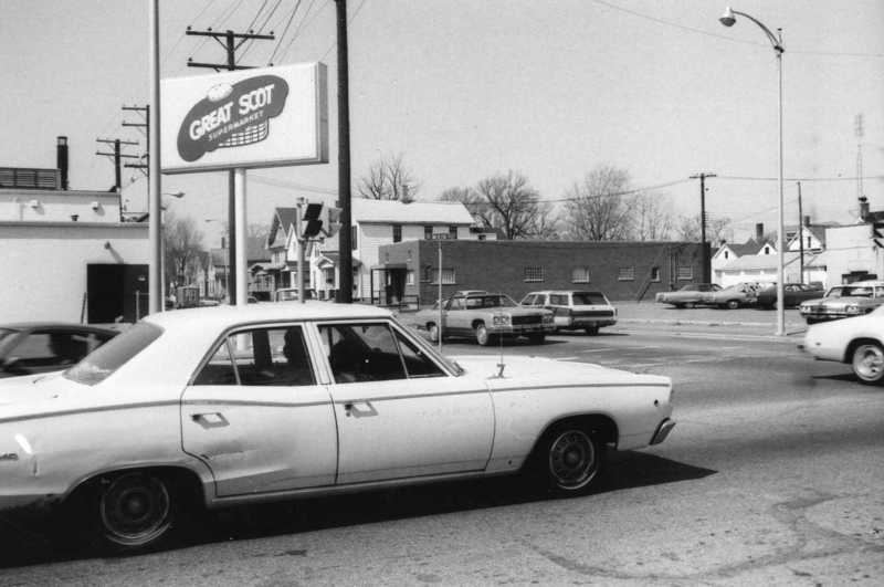 A vintage black and white photo of a street scene with old cars and a Great Scott billboard.