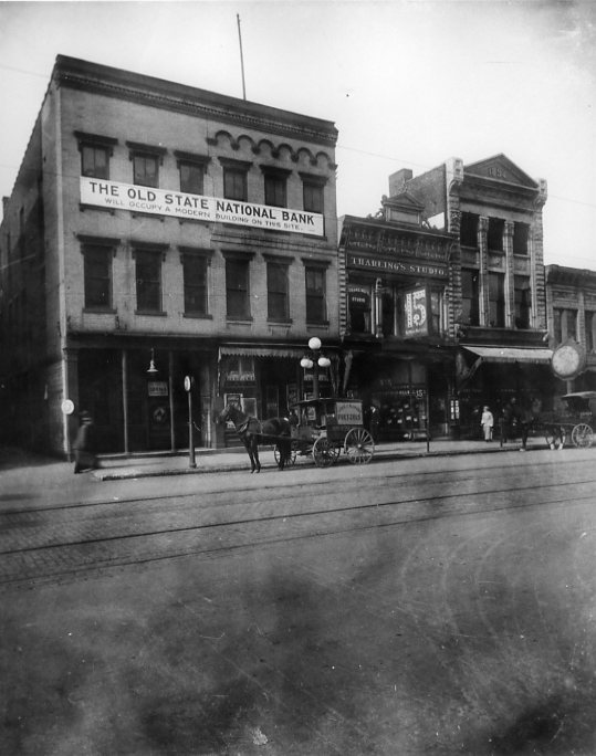 A historic black and white photo of building facades with "The Old State National Bank" sign, a horse-drawn carriage, and streetcar tracks.