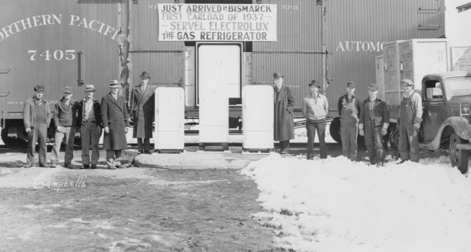 Vintage photo of men with new refrigerators by a train, snow on the ground, indicating a historical shipment arrival.