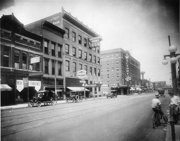 Vintage street scene with old cars, buildings with bank signs, and a person on a bike.