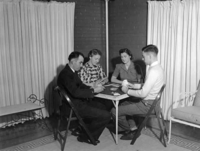 Four people are sitting around a small table, seemingly engaged in a card game or discussion, in a room with curtains.
