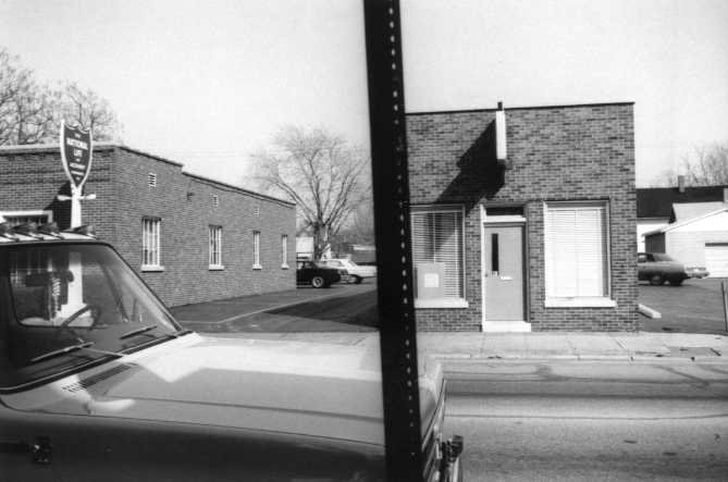 Black-and-white photo: Part of a car on the left, street view with buildings, and visible film edge.