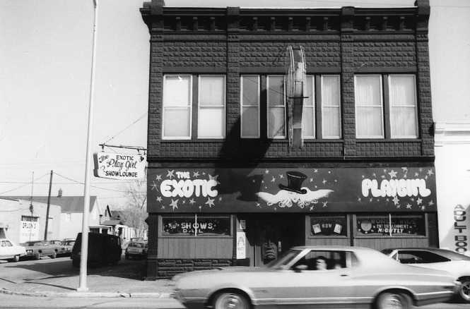 A black and white photo of a two-story building with signs for an exotic show lounge, a passing car in front.