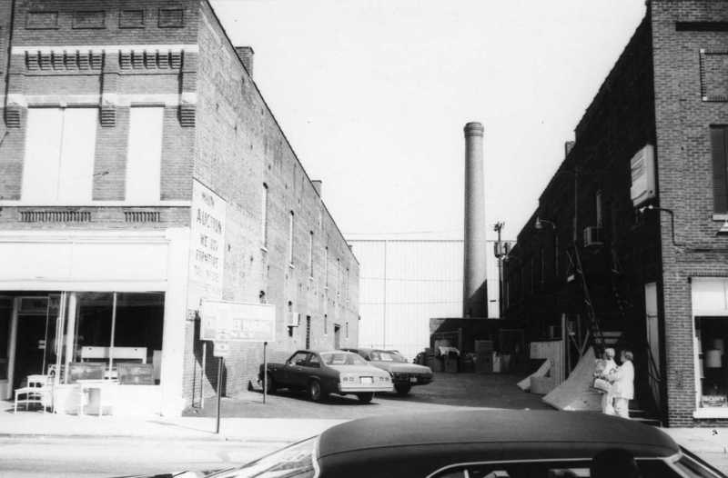 Black-and-white photo of an urban alley with old cars, buildings, and people chatting near stairs.
