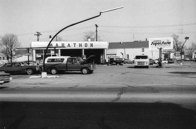 Monochrome photo of a vintage service station with parked cars and a street in the foreground.