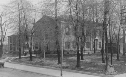 Black and white photo of a vintage two-story building behind bare trees, a sidewalk, and a classic car.