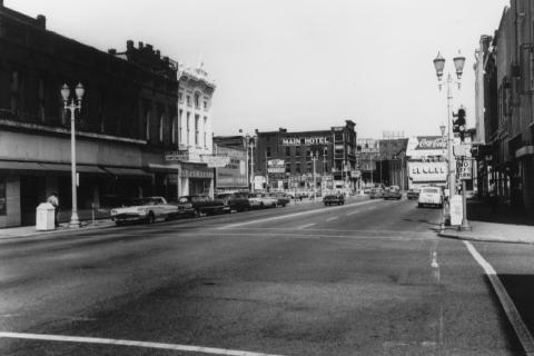 A vintage black and white photo of a main street with old cars, signage, buildings, and streetlights.