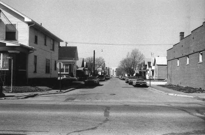 A black and white photo of a residential street with houses and parked cars, seemingly from an earlier era.