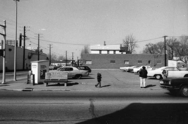 Street view with pedestrians, cars, a building, and a billboard. Black and white photo.