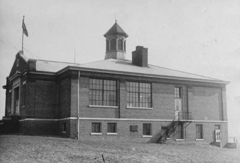 Old brick school building with large windows, a cupola, and an American flag.