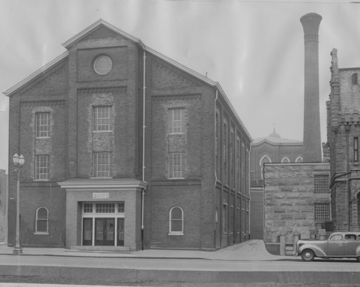 An old industrial brick building with large windows, a prominent entrance, and a vintage car parked beside it.