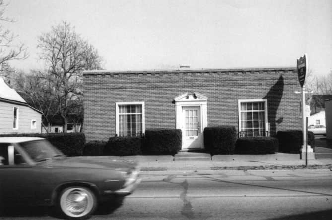 A blurred car passing in front of a brick building with a classic doorway and two windows, under a clear sky.