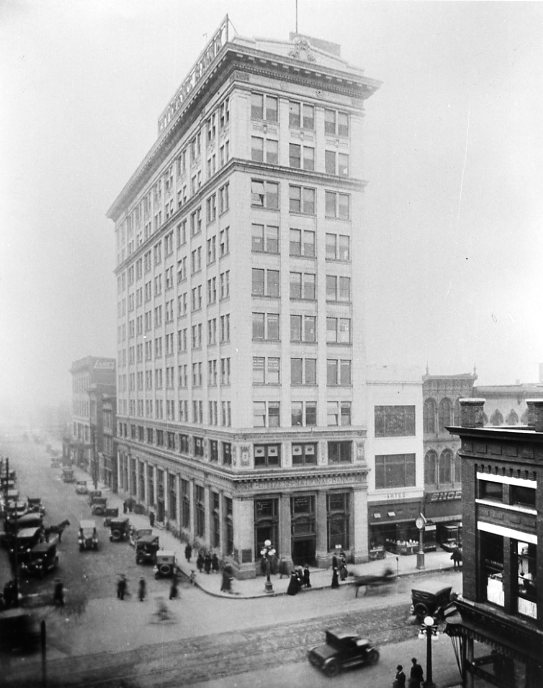 Vintage photo of a tall building on a city corner with pedestrians and old cars, enveloped in fog or smog.