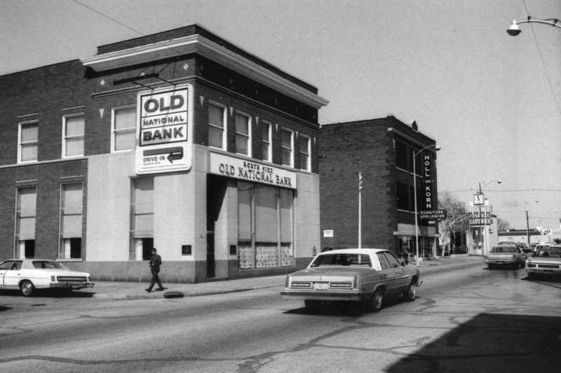 An old street with vintage cars, a person walking, and "Old National Bank" signage on a building.