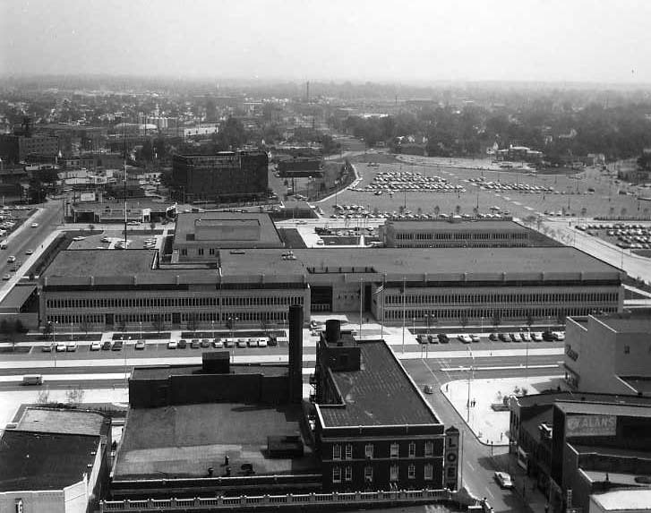 Black and white photo of a mid-20th-century cityscape with buildings, streets, and a large parking area. No people discernible.