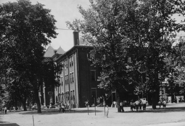 Historic photo of a brick building with large windows, surrounded by trees and people gathered outside, possibly a school or college.