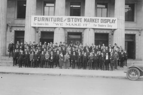A group of people posing in front of a building with a "Furniture & Stove Market Display" banner.