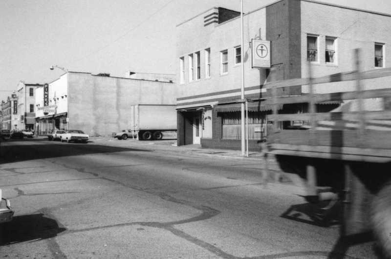 A black and white photo of a street with buildings, a parked semi-truck, cars, and a blurred foreground object.