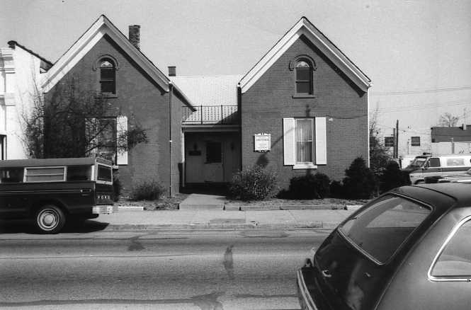 Two brick houses with gabled roofs, parked cars, and a "for sale" sign, in black and white.