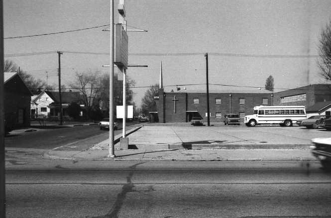 Black and white street view with buildings, a school bus, utility pole, and moving car.