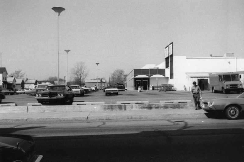 Parking lot with cars, buildings, a person, streetlights, and clear sky.