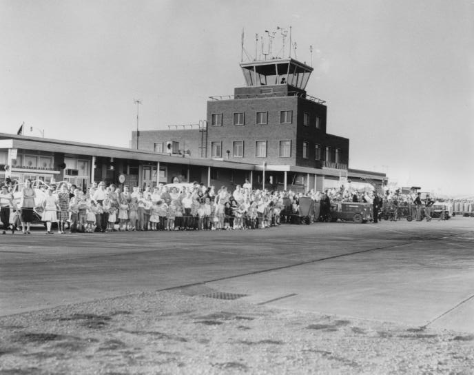 Black and white photo of a crowd gathered at an old airport with a control tower in the background.