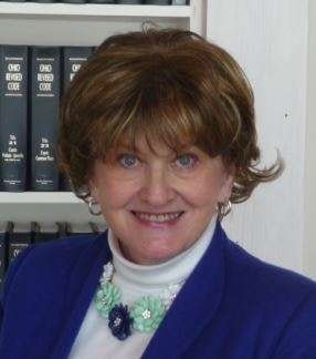 A smiling person with a blue jacket, white shirt, and earrings, standing in front of a bookshelf.