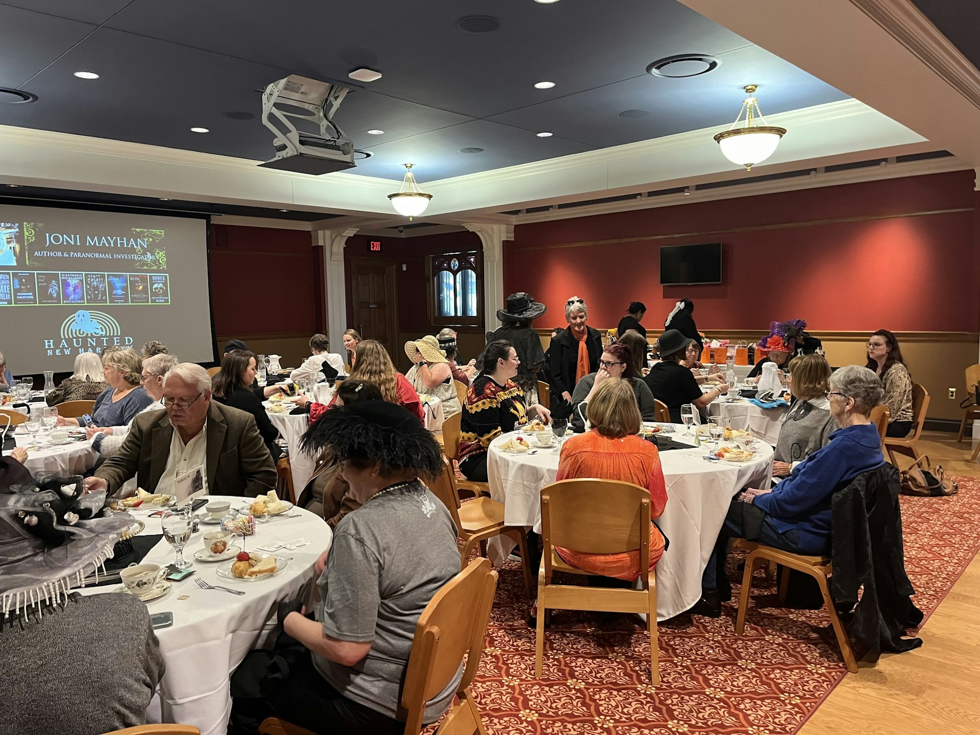 A group of people dressed in various styles dining at a banquet with a presentation on paranormal topics in the background.