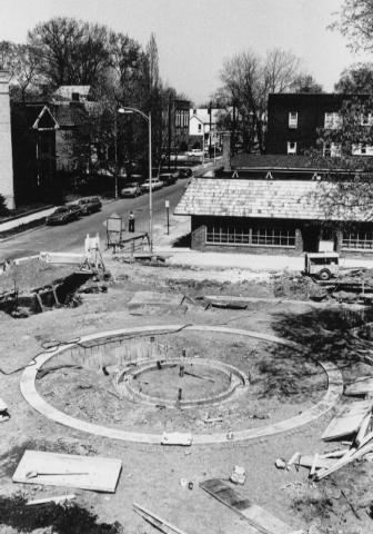 Circular foundation of a structure under construction with surrounding buildings and a street.
