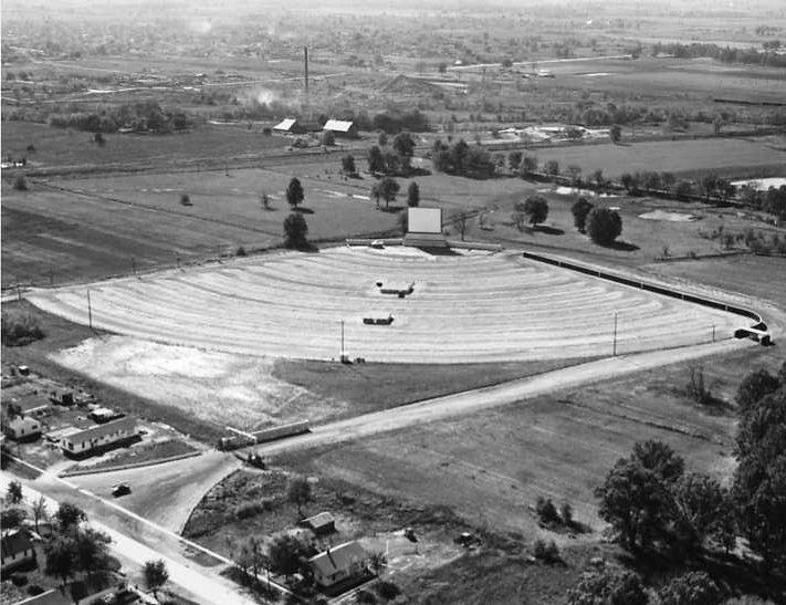 Aerial view of a classic drive-in movie theater with parked cars and large projection screen, amidst rural surroundings.