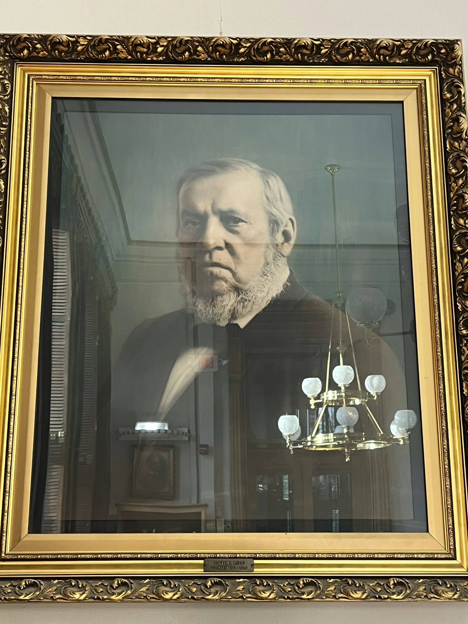 A framed portrait of an older man with a beard, hanging in a room. Reflections of a chandelier and other frames are visible.