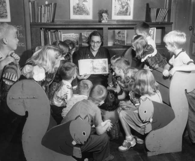 A group of children gather around an adult reading a book; some seats have whimsical animal designs. Black and white photo.
