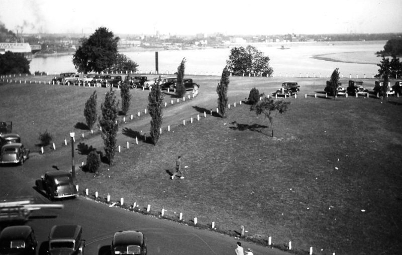 Vintage cars parked by a river, people walking on a grassy area with trees, and industrial smoke in the background.