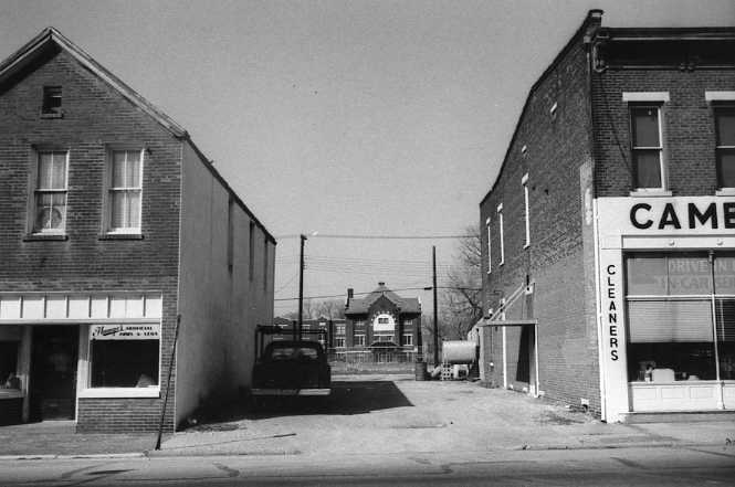 A black-and-white photo of an alley with older buildings, a parked truck, and a sign for a cleaners business.
