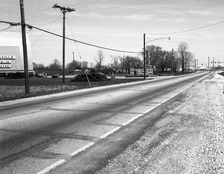 A black and white photo of a road with power lines and signs, including a drive-in theatre marquee. No people visible.