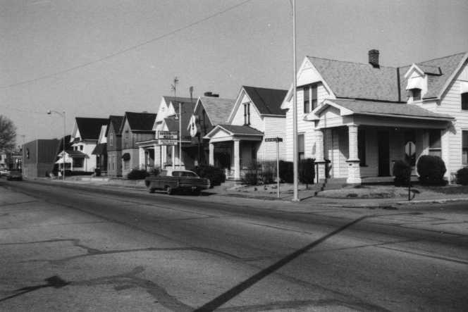 A black and white photo of a residential street with houses and a parked car.