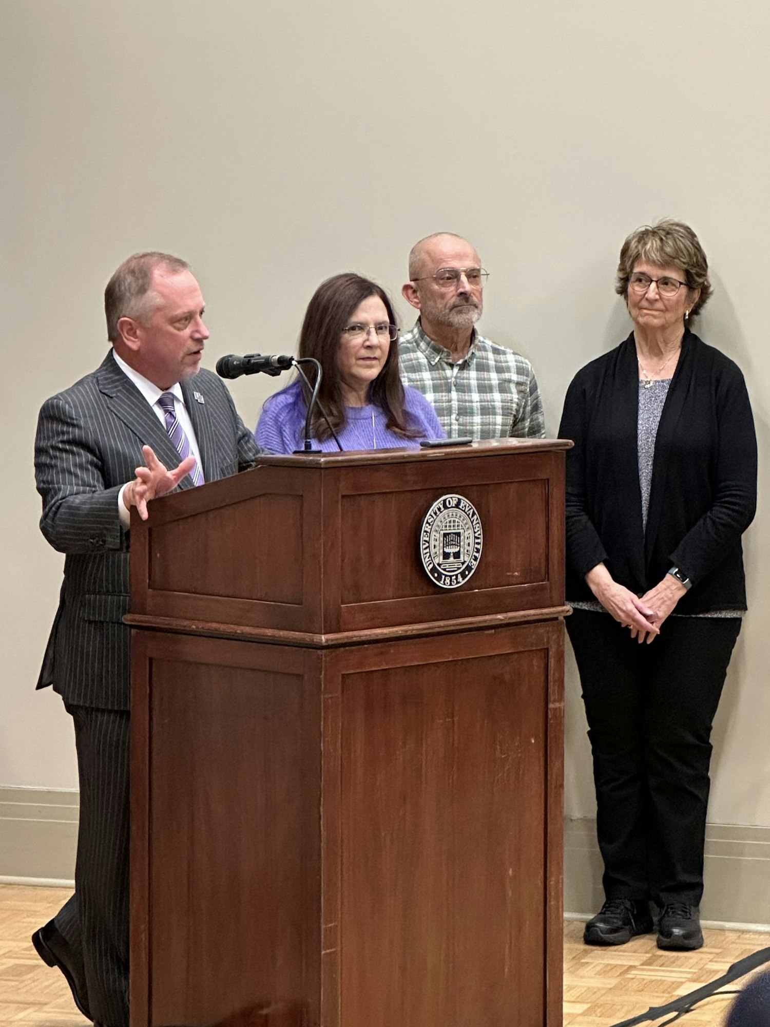 Man speaking at a podium with three attentive listeners behind him.