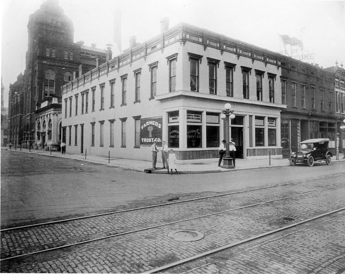 Historic street corner with a building, tram tracks, early car, and people, resembling early 20th-century city life.