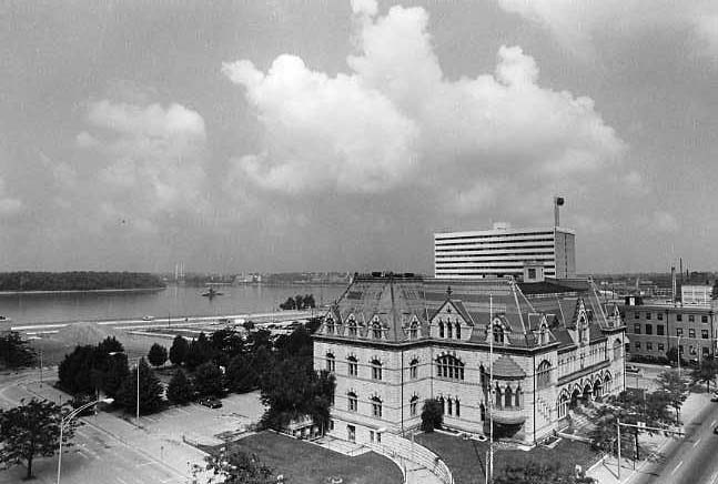 A black and white photo featuring a historical building in the foreground and a modern building behind it, near a body of water.