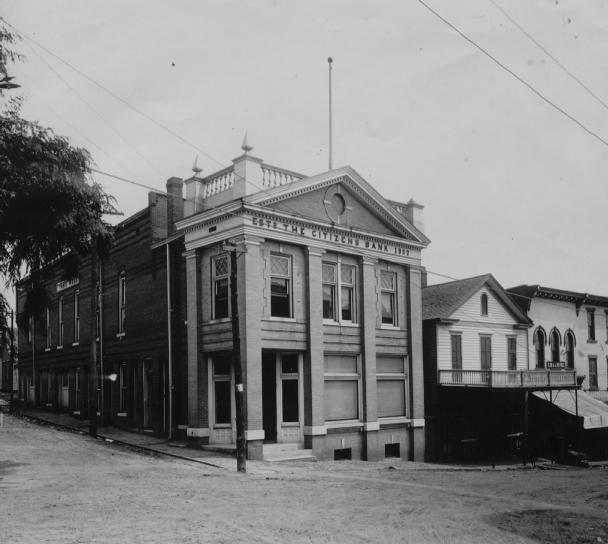 Vintage black and white photo of a classical two-story corner building with a sign "Bank" and adjacent houses.