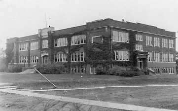 Old two-story building with large windows, likely a school or administrative structure, surrounded by a grassy lawn and a sidewalk.
