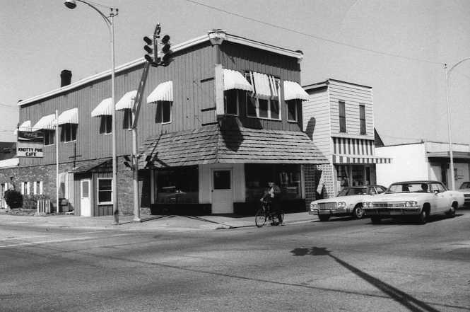 Black and white photo of a street corner with a vintage building, cars, and a cyclist.