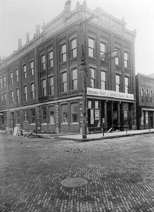 Vintage corner building with "INDIANA TRUST & SAVINGS BANK" sign, cobblestone street, no people visible.