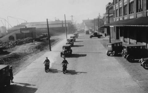 Old street with vintage cars and motorcycles, industrial buildings, possibly early 20th century.