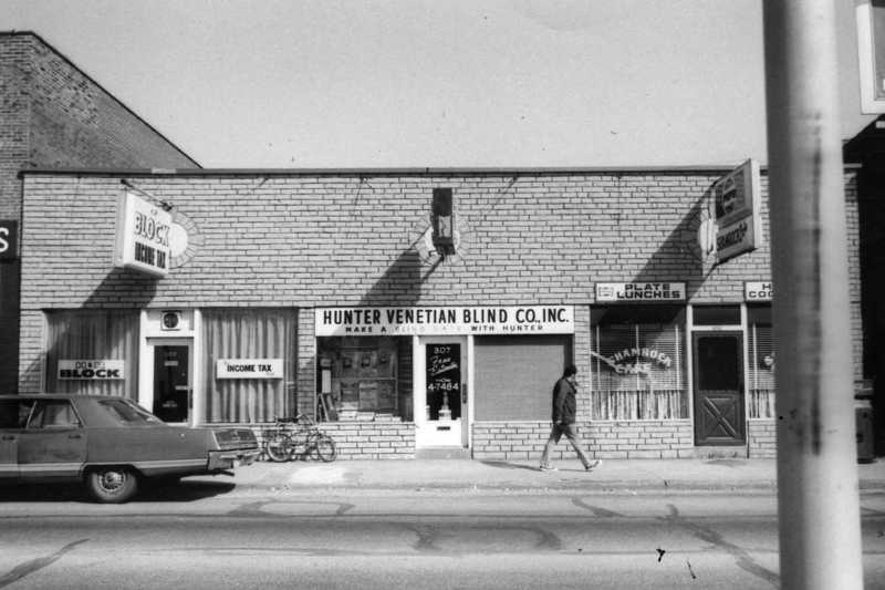 A vintage streetscape showing small businesses including "HUNTER VENETIAN BLIND CO. INC," a person walking, parked cars, and bicycles.