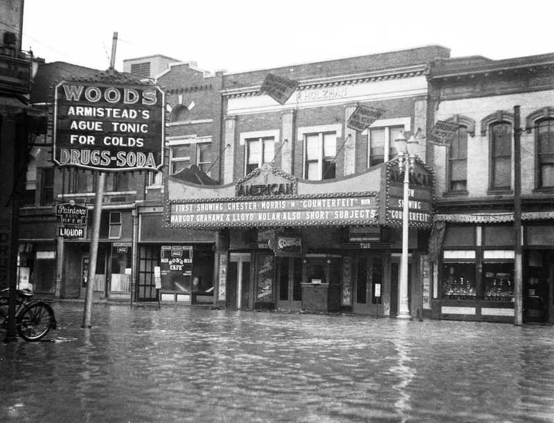 An old flooded street with submerged storefronts, including a pharmacy advertising drugs and soda, and a theater marquee.