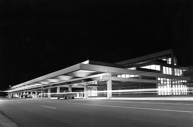 Modern building at night with lit interior, long overhangs, and parked vehicles. Black-and-white photo.