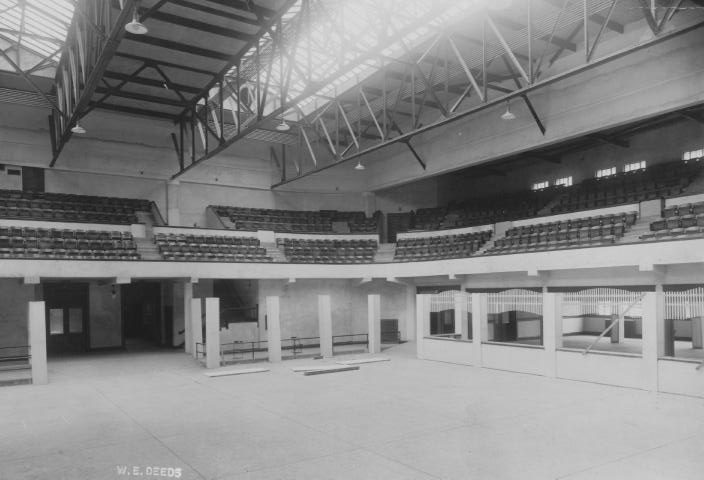 Empty indoor arena with tiered seating and overhead rafters, possibly historic.