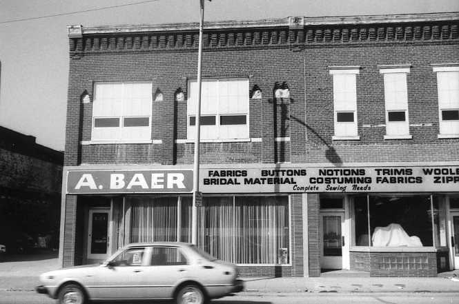 Black and white photo of a vintage fabric store front with a car passing by.