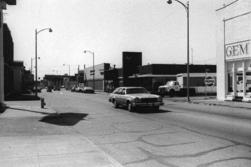 A black-and-white photo of a vintage street scene with old cars, buildings, and a Pepsi sign.